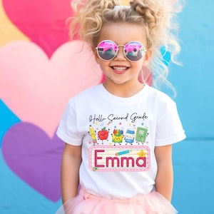 Pu&ograve; includere: Una t-shirt bianca a maniche corte con una grafica colorata che riporta il testo Hello Second Grade e il nome Emma. Il design include illustrazioni a cartone animato di una matita, una mela, pastelli, un cartone di latte, un quaderno e una stella.