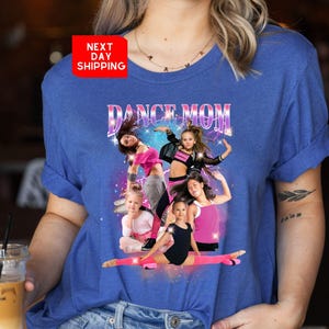 K&ouml;nnte beinhalten: Ein blaues T-Shirt mit dem Text "Dance Mom" in rosa und lila Buchstaben. Das Shirt zeigt eine Grafik mit f&uuml;nf M&auml;dchen in Tanzposen vor einem glitzernden Hintergrund.