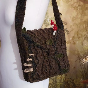K&ouml;nnte beinhalten: Eine braune geh&auml;kelte Umh&auml;ngetasche mit Baumrindenmuster. Die Tasche hat einen Riemen und ist mit kleinen geh&auml;kelten Pilzen verziert.