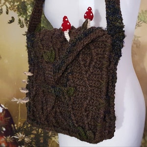 K&ouml;nnte beinhalten: Eine braune geh&auml;kelte Tasche mit einem Blattdesign und zwei roten und wei&szlig;en Pilzverzierungen. Die Tasche hat einen langen Riemen und wird an einem wei&szlig;en Mannequin getragen.
