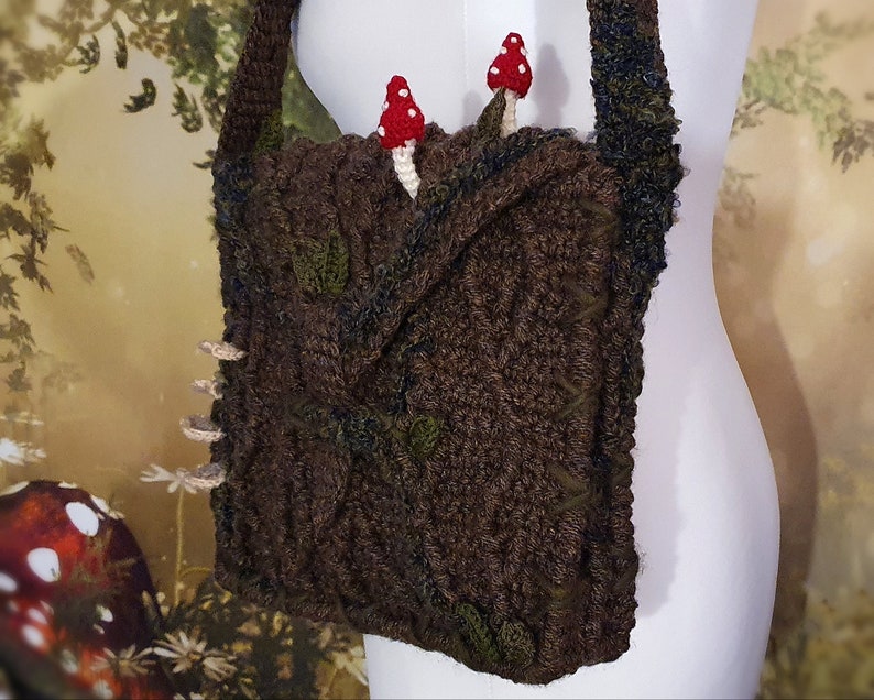 K&ouml;nnte beinhalten: Eine braune geh&auml;kelte Tasche mit einem Blattdesign und zwei roten und wei&szlig;en Pilzverzierungen. Die Tasche hat einen langen Riemen und wird an einem wei&szlig;en Mannequin getragen.