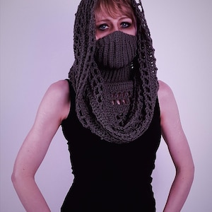 May include: A dark gray crocheted hood and cowl set. The hood features an open, lacy design around the face and head, while the cowl covers the neck and lower face. The person is wearing a black tank top.