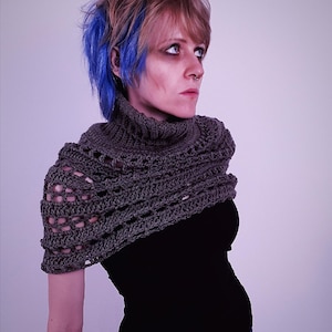 May include: A gray crocheted shawl with a high collar. The shawl is made of a textured yarn and has an open, lacy design. The person is wearing a black top and has blue and blonde hair.