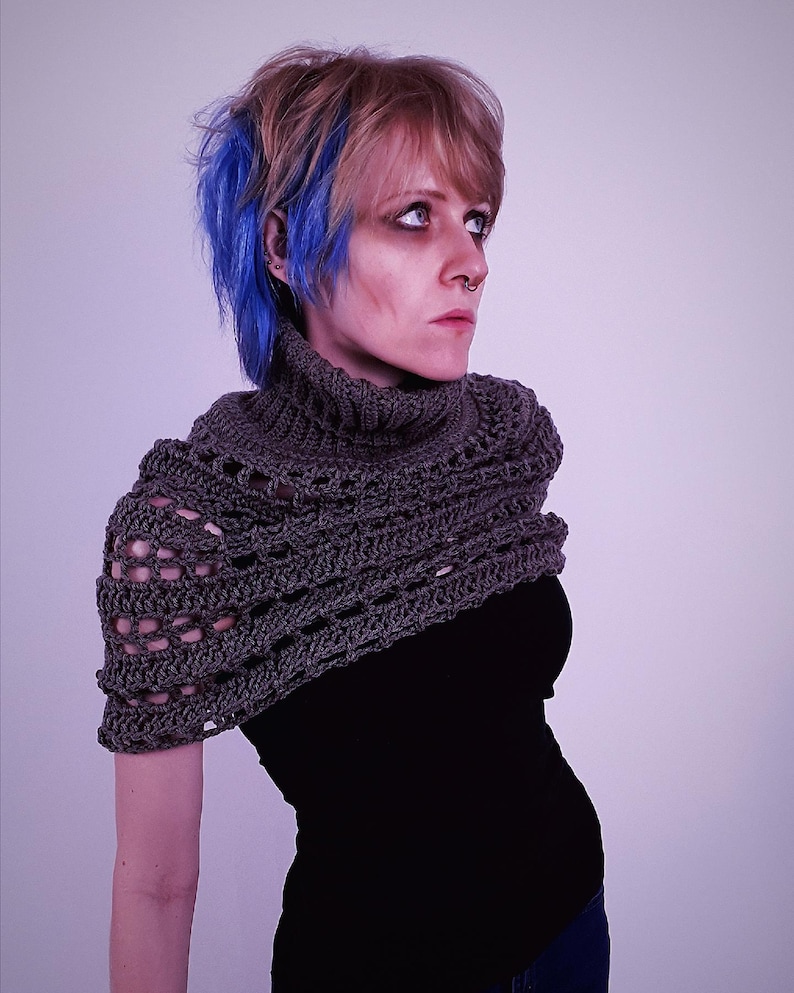 May include: A gray crocheted shawl with a high collar. The shawl is made of a textured yarn and has an open, lacy design. The person is wearing a black top and has blue and blonde hair.
