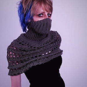 May include: A gray crocheted cowl and shoulder wrap. The cowl covers the lower face, and the wrap features an open, lacy design. The garment is worn over a black top. The model has blue streaks in their hair.