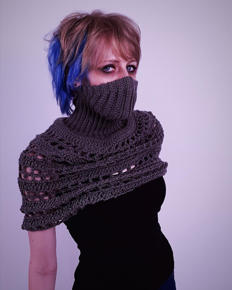 May include: A gray crocheted cowl and shoulder wrap. The cowl covers the lower face, and the wrap features an open, lacy design. The garment is worn over a black top. The model has blue streaks in their hair.