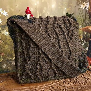 K&ouml;nnte beinhalten: Eine braune geh&auml;kelte Tasche mit Waldmotiv. Die Tasche hat einen Klappverschluss und einen kleinen roten Pilz oben. Die Tasche liegt auf einer Holzoberfl&auml;che mit aufwendigen Schnitzereien.