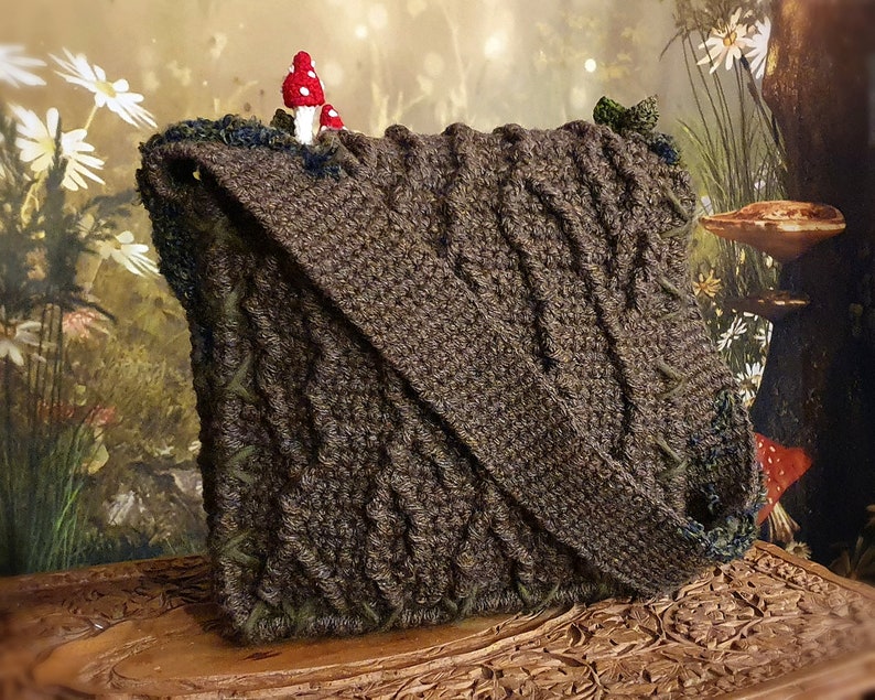 K&ouml;nnte beinhalten: Eine braune geh&auml;kelte Tasche mit Waldmotiv. Die Tasche hat einen Klappverschluss und einen kleinen roten Pilz oben. Die Tasche liegt auf einer Holzoberfl&auml;che mit aufwendigen Schnitzereien.
