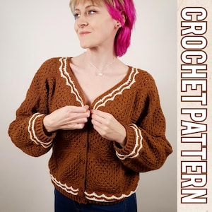May include: A brown crochet cardigan with white trim. The cardigan has a V-neck and button closure. The model is wearing the cardigan with blue jeans.