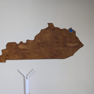 Könnte beinhalten: Ein Holzausschnitt des Bundesstaates Kentucky, braun gebeizt, mit einem blauen Stern-Aufkleber an der Stelle von Lexington.