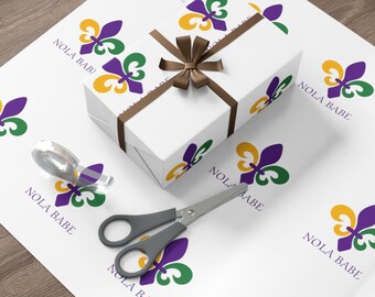 Puede incluir: Papel de regalo blanco con un patrón repetido de flores de lis moradas y doradas con el texto "NOLA BABE" impreso en cada una. Un lazo marrón está atado en la parte superior de una caja de regalo blanca envuelta en el mismo papel.
