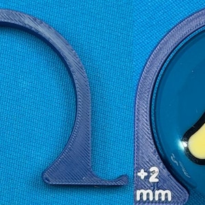 May include: A blue, 3D-printed semi-circular object with a flat base and a measurement of +2 mm. The object is next to a blue ball with a yellow triangle and the number 2 inside. The ball is also surrounded by a blue frame.