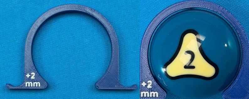 May include: A blue, 3D-printed semi-circular object with a flat base and a measurement of +2 mm. The object is next to a blue ball with a yellow triangle and the number 2 inside. The ball is also surrounded by a blue frame.