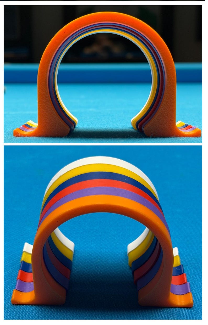 May include: A colorful, arched pool table accessory. The object is orange with layers of yellow, blue, purple, and red. The accessory is designed to hold pool balls.