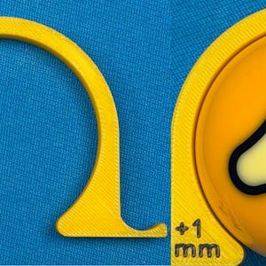 May include: A yellow 3D-printed tool with a curved design, featuring a measurement of +1 mm. The tool is positioned next to a yellow billiard ball with a black and white graphic of the number 1.