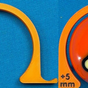 May include: An orange pool ball with the number 5 inside a white and black triangle, next to an orange curved guide. The guide has "+5 mm" printed on it. The background is blue.