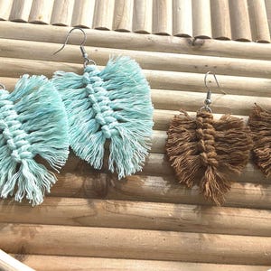 Puede incluir: Dos pares de pendientes de macramé hechos a mano. Un par es azul claro, el otro marrón. Los pendientes tienen forma de hoja con un centro anudado y bordes con flecos, colgando de ganchos plateados. Los pendientes se exhiben sobre una superficie de madera.