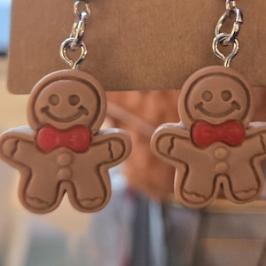 May include: A pair of dangle earrings featuring light brown gingerbread man charms. Each charm has a smiling face, a bright red bow tie, and two small buttons on the chest. The charms are attached to silver-toned metal earring hooks.