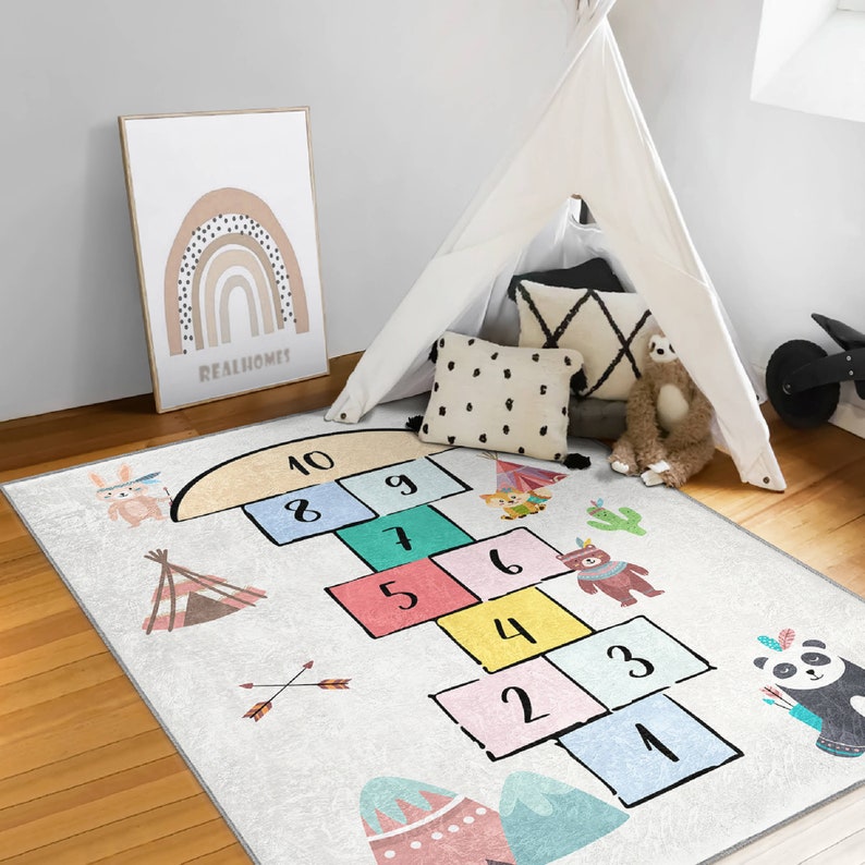 Op de afbeelding: Een wit en grijs speelkleed met een tikkertje-spel met kleurrijke vierkanten genummerd van 1 tot 10. Het kleed heeft ook een verscheidenheid aan andere ontwerpen, waaronder pijlen, tipi's en dieren.  Realhomes is afgedrukt op een ingelijst beeld op de achtergrond.