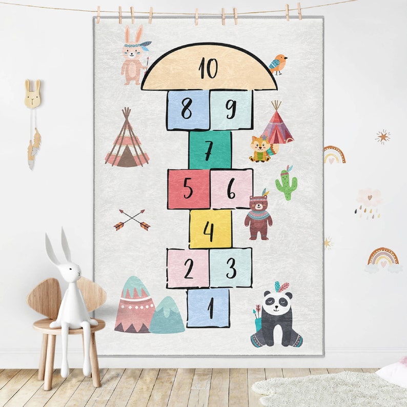 Op de afbeelding: Een kleurrijk hinkelspelkleed met een sprookjesachtig bosmotief. Het kleed heeft de nummers 1-10, een tipi, een cactus, een vos, een beer, een panda, een konijn en een vogel.
