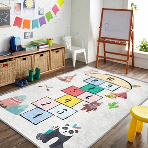 Op de afbeelding: Een wit speelkleed met een hopscotch-ontwerp met kleurrijke vierkanten genummerd van 1 tot 10. Het kleed heeft ook een pandabeer, een beer en een cactus.
