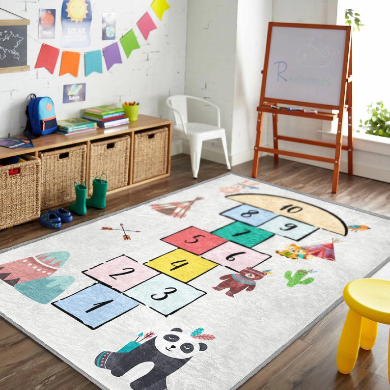 Op de afbeelding: Een wit speelkleed met een hopscotch-ontwerp met kleurrijke vierkanten genummerd van 1 tot 10. Het kleed heeft ook een pandabeer, een beer en een cactus.