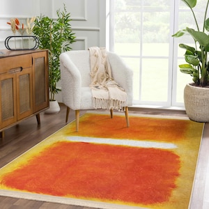 May include: A white armchair with a cream throw blanket sits on a yellow, orange, and white abstract area rug. The rug has a white stripe in the middle.
