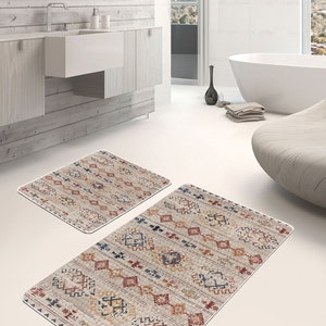 Op de afbeelding: Twee badmatten met patronen in een badkamer. De matten zijn beige met rode, blauwe en gele geometrische patronen. De matten liggen op een witte tegelvloer.