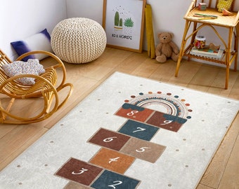 Puede incluir: Alfombra rectangular blanquecina con diseño de rayuela. Los cuadrados de la rayuela son en tonos marrón, azul y naranja, numerados del 1 al 9. Un arcoíris decorativo y un patrón de puntos en la parte superior. La alfombra está en una habitación infantil.
