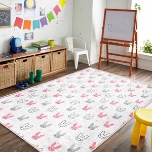 May include: A white and pink rug with a repeating pattern of grey and pink bunny faces. The rug is on a wooden floor in a room with a white brick wall, a wooden shelf with baskets, and a white easel with a chalkboard.
