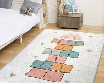 May include: A rectangular children's rug featuring a hopscotch design. The rug has a light gray background with a hopscotch grid in various colors, including pink, teal, orange, and gray. A rainbow and hearts decorate the top of the rug.