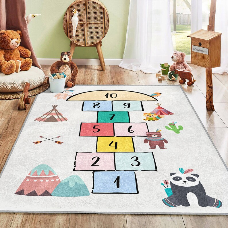 Op de afbeelding: Een wit en grijs speelkleed met een hinkelbaanontwerp met de nummers 1 tot en met 10. Het kleed heeft een kleurrijk ontwerp met dieren en een tipi.