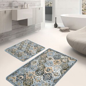 Op de afbeelding: Twee badmatten met een geometrisch patroon in blauw, beige en bruin. De matten liggen op een witte tegelvloer in een badkamer.