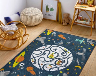 May include: A blue and yellow space-themed rug with a large circle in the center depicting a planet with roads and buildings. The rug features stars, planets, rockets, and other space-related imagery.