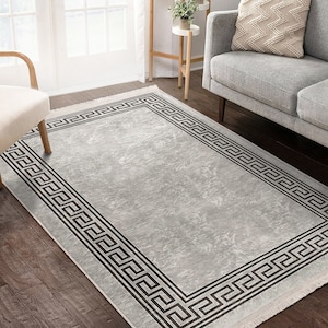 Puede incluir: Una alfombra gris claro con un borde negro y blanco de clave griega. La alfombra tiene un aspecto desgastado y un borde de flecos.