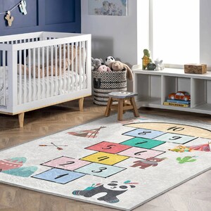 Op de afbeelding: Een wit en grijs speelkleed met een hinkelbaanontwerp met de nummers 1 tot en met 10. Het kleed heeft ook illustraties van een pandabeer, een tipi en cactussen.