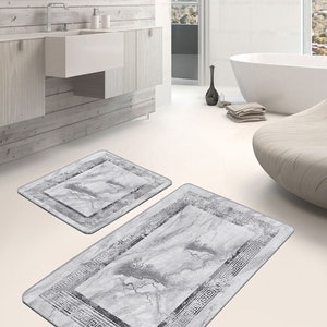 Puede incluir: Dos alfombras de baño grises con un patrón de mármol y un borde geométrico negro. Las alfombras están sobre un suelo de baldosas blancas en un baño.