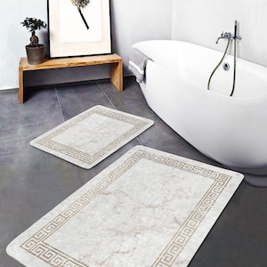 Pode incluir: Dois tapetes de banho brancos com uma borda bege de chave grega. Os tapetes têm um padrão de mármore. Os tapetes estão colocados num chão de azulejos cinzentos numa casa de banho.