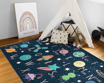 May include: A blue alphabet rug with colorful illustrations of planets, stars, rockets, and aliens. The letters are in a circular pattern around the edge of the rug. The rug is on a wooden floor in a room with a teepee tent and a rainbow picture.