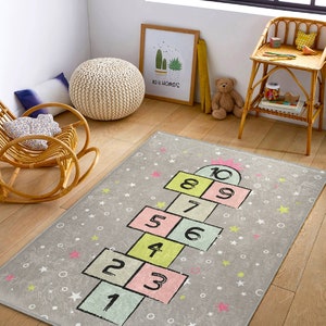 Könnte beinhalten: Ein grauer und weißer Hopscotch-Teppich mit rosa, grünen und gelben Quadraten, die von 1 bis 10 nummeriert sind. Der Teppich hat einen weißen Hintergrund mit grauen Sternen und weißen Kreisen.