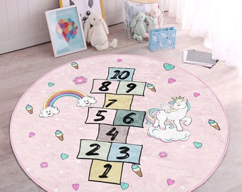 May include: A pink round rug with a hopscotch game, a rainbow, clouds, ice cream cones, and a unicorn with a crown and wings. The rug has a pink background with white stars and pink hearts.