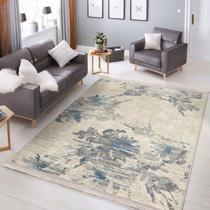 May include: A light beige area rug with a blue and gray floral pattern. The rug has a fringe border and is placed in a living room setting with a gray sofa and armchair.