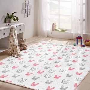 May include: A white and pink children's rug with a repeating pattern of gray bunny faces with pink ears and noses. The rug has a pink border.