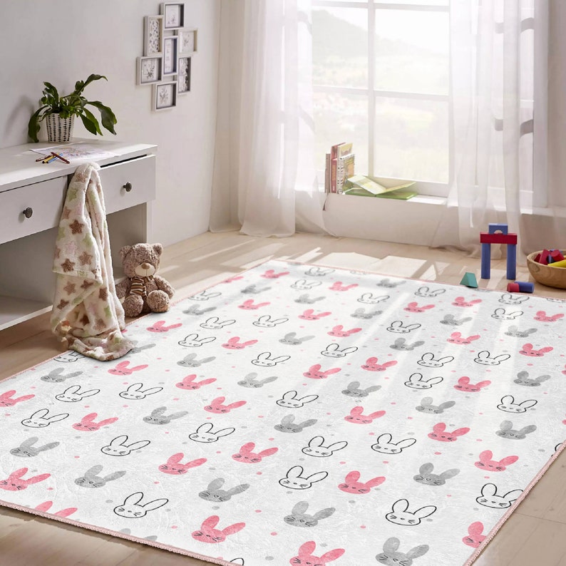 May include: A white and pink children's rug with a repeating pattern of gray bunny faces with pink ears and noses. The rug has a pink border.