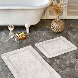 Puede incluir: Dos alfombras de baño beige con un diseño de llave griega. Las alfombras están sobre un suelo de baldosas grises en un baño.