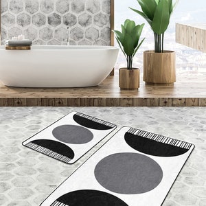 Könnte beinhalten: Zwei weiße Badematten mit schwarzen und grauen geometrischen Mustern. Die größere Matte hat einen großen grauen Kreis mit einem schwarzen Halbkreis oben. Die kleinere Matte hat einen kleineren grauen Kreis mit einem schwarzen Halbkreis oben.