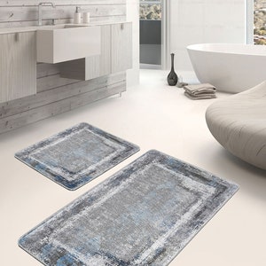 Op de afbeelding: Twee badmatten in een moderne badkameromgeving. De matten zijn rechthoekig en hebben een verweerd, abstract ontwerp in grijstinten en blauw. De grotere mat bevindt zich op de voorgrond, terwijl de kleinere mat gedeeltelijk zichtbaar is op de achtergrond.