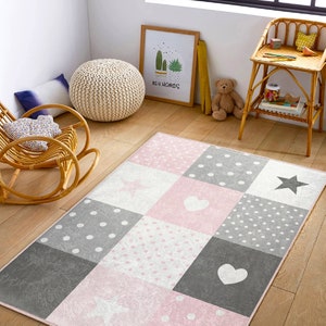May include: A pink and gray patchwork rug with white polka dots, stars, and hearts. The rug is on a wooden floor in a room with a rocking chair and a small table.