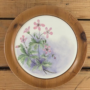 Pode incluir: Um prato redondo com moldura de madeira, fundo branco e um desenho pintado de flores cor-de-rosa e folhas verdes. As flores são pintadas em estilo aquarela e têm um aspeto suave e delicado.