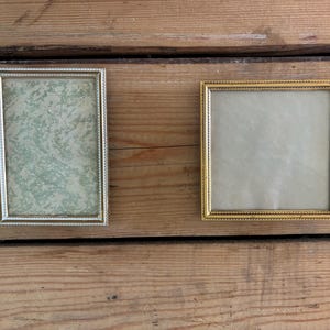 Può includere: Due cornici vintage. Una è rettangolare con un bordo argentato perlato e un interno a fantasia verde e bianco. L'altra è quadrata con un bordo dorato perlato e un interno color crema. Entrambe sono vuote.